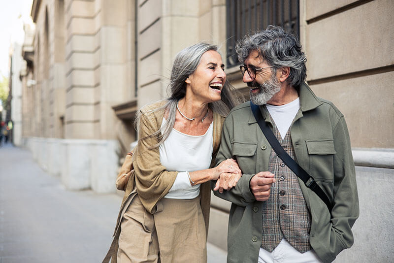 An older couple walks arm in arm down a city street, smiling and laughing despite the mans hearing loss. The woman, with long gray hair, wears a light outfit, while the man sports glasses and a green jacket. Theyre enjoying their joyful conversation.