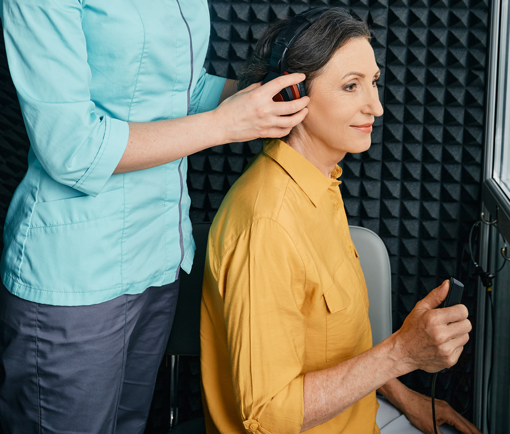 An audiologist places headphones on an older woman in a soundproof room during a hearing test. The woman is holding a response button and smiling slightly.