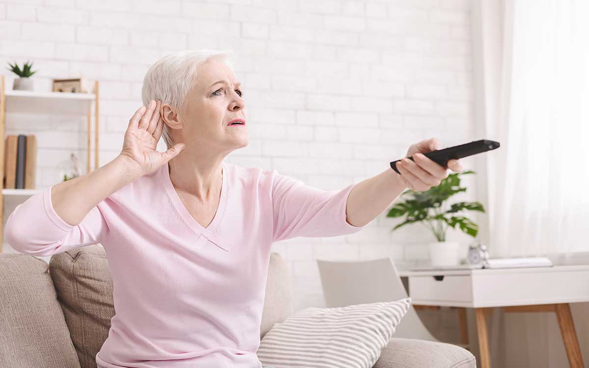 Older woman with short white hair sits on a couch, holding a TV remote and cupping her ear as if struggling to hear, in a bright living room with white brick walls and modern decor.