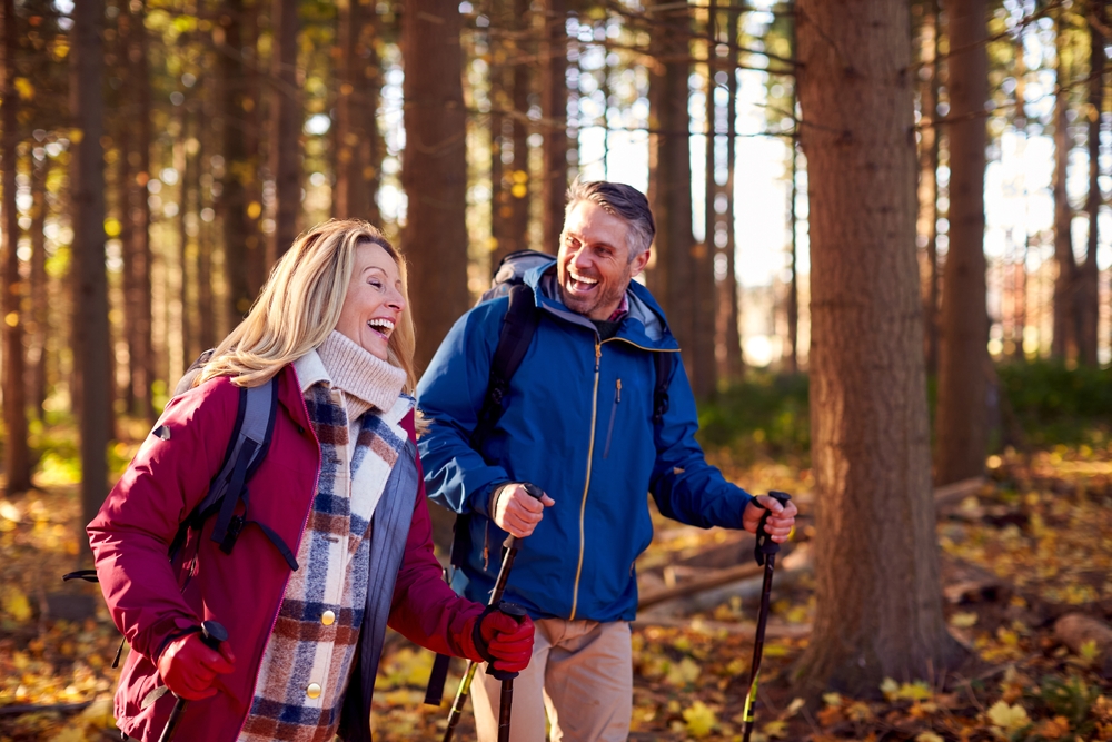 A smiling man and woman wearing jackets and backpacks walk through a sunlit forest with hiking poles. Fallen leaves cover the ground, and tall trees surround them, suggesting an autumn hike.