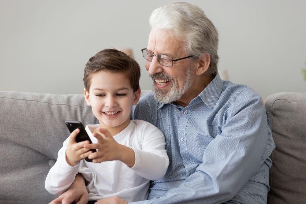 A smiling elderly man with glasses sits on a couch with a young boy. They are both looking at and holding smartphones, appearing happy and engaged.
