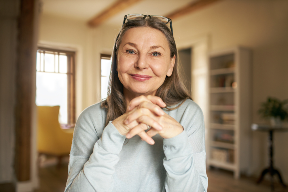 A smiling older woman with long gray hair and glasses resting on her head sits indoors with her hands clasped, wearing a light blue sweater. The background shows a bright, cozy room with shelves and a window.
