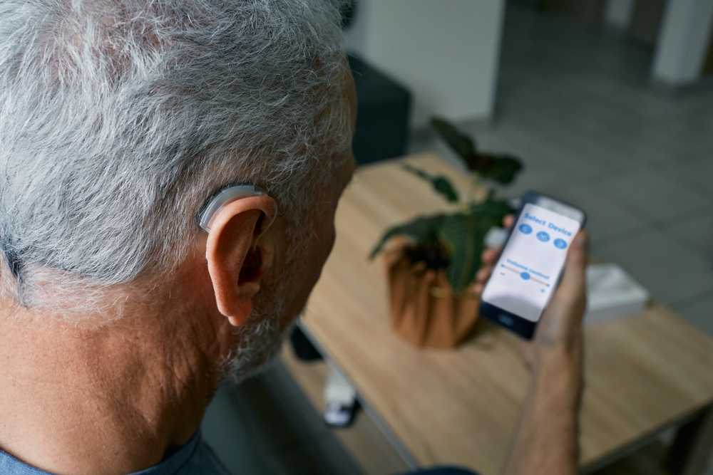 An older man with gray hair and a hearing aid looks at a smartphone displaying an app, sitting at a wooden table with a potted plant in the background.