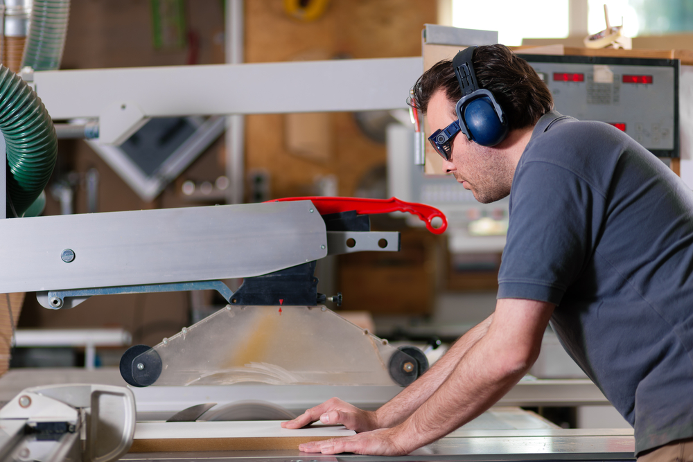 A man wearing safety glasses and earmuffs operates a large table saw in a workshop. He is focused on guiding a wooden board through the saw.