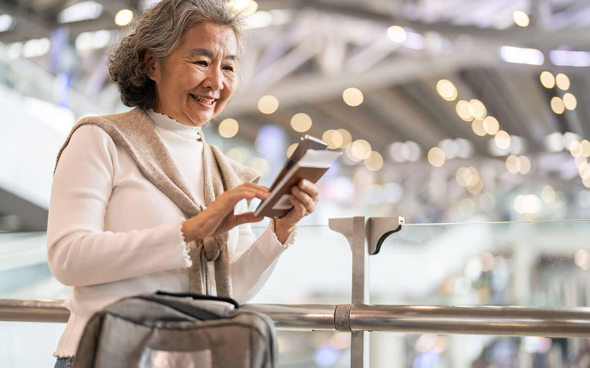 An older woman smiles while using her smartphone and holding a passport at an airport. She stands near a railing with a bag and blurred lights in the background, suggesting she is ready to travel.