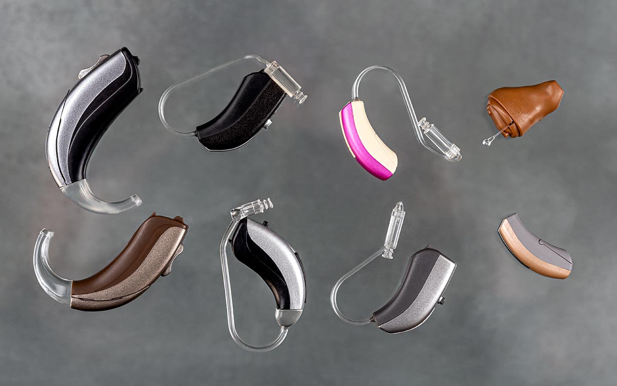 A group of eight hearing aids in various shapes, sizes, and colors arranged on a gray background. Some have flesh-toned, black, silver, or pink accents, and feature clear plastic ear tubes or molds.